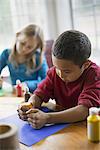 Children in a family home.  Two children sitting at the table, using paint and paper to create decorations.