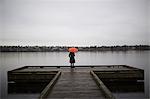 A woman wearing all black and holding an orange umbrella stands on the edge of a dock on a grey and cloudy day in Seattle, WA