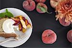 A table viewed from overhead. Organic fruit, peaches, and flowers. a plate with fresh fruit, cake and creme fraiche. A fork. Dessert.