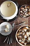 A cheese fondue on a table top. Cubed bread and long forks, for dipping bread into rich melted cheese mixture.