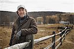 A man leaning against a post and rail fence on a farm in winter.