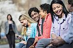 People outdoors in the city in spring time. Five people sitting in a row, one looking at a mobile phone. A woman approaching in the distance.