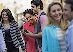 People outdoors in the city in spring time. A group of men and women, two looking at a cell phone screen and laughing.