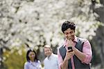 People outdoors in the city in spring time. Cherry blossom on the trees. A man checking his cell phone, and two people behind him.