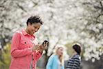 People outdoors in the city in spring time. White blossom on the trees. A young woman checking her cell phone, and laughing.