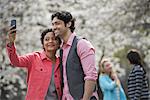 People outdoors in the city in spring time. White blossom on the trees. A young woman taking a photograph of herself and a young man.