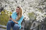 Outdoors in the city in spring time. New York City park. White blossom on the trees. A woman sitting on a bench holding her mobile phone.