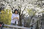 Outdoors in the city in spring time. New York City park. White blossom on the trees. A woman holding her mobile phone and smiling.