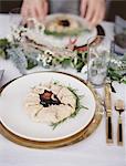 A table top covered with  a white cloth. A place setting with cutlery and plate. A green foliage table decoration. A meal set on a plate.