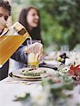 A group of people around a table in a garden. A celebration meal, with table settings and leafy decorations. A person pouring drinks into glasses.