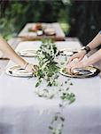 Two people leaning over a table laid outside with a white cloth and a central foliage table decoration. Placing cutlery and plates on a tabletop.