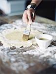 A domestic kitchen. A woman preparing a meal. Rolling out pastry on a table top and brushing it with egg wash.