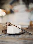 A domestic kitchen. A woman in the background. A slab of fresh organic butter, on a wooden dish.