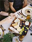 A table laid with a white cloth and place settings seen from above. An organic cheese board with soft and hard cheeses and figs. Two people sitting at the table.