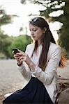 A woman wearing a cream jacket, sitting in a city park looking at her cell phone.