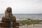 A woman with long blonde hair wearing a brown hooded coat, seated on a bench looking out to sea.