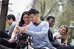 City life in spring. Young people outdoors in a city park. Sitting on a bench. A man showing his phone screen to a woman, and three other people checking their phones.