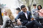 City life in spring. Young people outdoors in a city park. A group of men and women gathered around a park bench. Two checking a smart phone for messages.