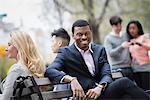 City life in spring. Young people outdoors in a city park. A group of men and women gathered around a park bench. Two checking a smart phone for messages.