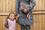 A woman in a grey coat holding a black and white chicken with a red coxcomb under one arm. A young girl beside her holding her other hand.