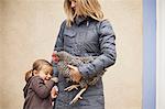 A woman in a grey coat holding a black and white chicken with a red coxcomb under one arm. A young girl beside her holding her other hand