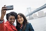 New York city. The Brooklyn Bridge crossing over the East River. A couple, two women, taking a picture with a phone, a selfy of themselves.