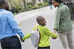 A New York city park in the spring. A boy with a bookbag, holding hands with his mother and father.