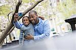 A New York city park in the spring. Sunshine and cherry blossom. A father and son side by side. Using a smart phone to take a picture.