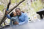 A New York city park in the spring. Sunshine and cherry blossom. A father and son side by side. Using a smart phone to take a picture.