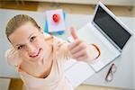 Happy young woman with books and laptop in kitchen showing thumbs up