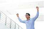 Successful Asian Indian businessman with arms up celebrating his victory, modern office building as background.