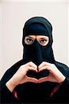 Close-up portrait of young woman wearing black, muslim hijab and muslim dress, making heart shape with hands, looking at camera, eyes showing eye makeup, studio shot on white background