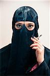 Close-up portrait of young woman wearing black, muslim hijab and muslim dress, holding cigarette and smoking, looking at camera, eyes showing eye makeup, studio shot on white background