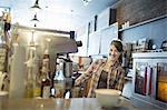 A person, barista, behind the counter at a coffee shop. A large coffee machine for making fresh coffee. A cappuccino in a white cup on the counter.