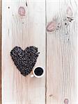 Overhead view of a heap of roasted organic coffee beans in a heart shape, arranged on a tabletop. A cup of black coffee.