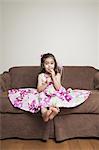 A 3 year old girl with long brown hair in a pink flowered cotton dress with the skirt spread out, sitting on a brown sofa.