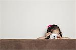 A young three year old girl crouching behind a sofa, with a toy camera, taking a picture.