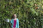A young girl standing in front of a wall covered with ferns and climbing plants.