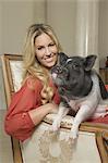 A miniature pot bellied pig sitting on his haunches on the lap of a blonde haired woman, sharing an antique armchair with thick cushions, in a large elegantly furnished mansion in Texas.