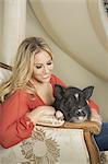 A miniature pot bellied pig sitting on his haunches on the lap of a blonde haired woman, sharing an antique armchair with thick cushions, in a large elegantly furnished mansion in Texas.