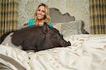 A pot bellied pig lying on a large bed with carved headboard and pillows, in a large mansion, beside the owner, a woman with blonde hair.