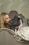 A pot bellied pig lying on a large bed with carved headboard with the owner, a blonde haired woman kissing her pet on the snout.