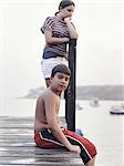 Two young people, teenagers, boy and girl, on a dock overlooking moored boats on the coastline.