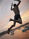 A boy leaping off the dock into the water, at sunset on the coast.