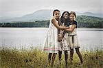 Three young girls standing by the side of a lake, hugging each other.