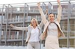 Excited young businesswomen with arms raised against office building