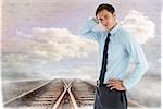 Thinking businessman with hand on head against train tracks leading to brick wall with clouds