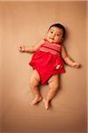 Portrait of Asian baby lying on back, wearing red dress, looking at camera and smiling, studio shot on brown background