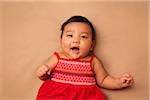 Close-up portrait of Asian baby lying on back, wearing red dress, looking at camera and smiling, studio shot on brown background