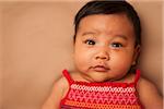 Close-up portrait of Asian baby lying on back, wearing red dress, looking at camera, studio shot on brown background